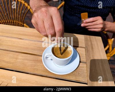 La main blanche d'un homme caucasien agite une tasse de café fraîchement brassée sur une terrasse sur une table en bois. Il n'existe aucune personne ou marque de commerce reconnaissable dans Banque D'Images