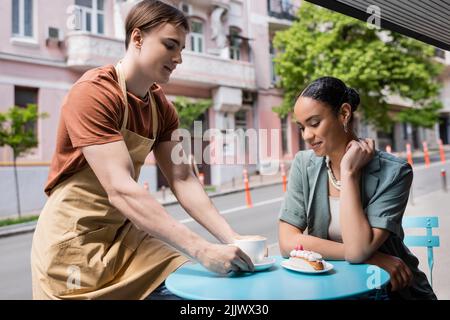 Vendeur en tablier mettant le cappuccino près du dessert et client afro-américain sur la terrasse de la sucrée Banque D'Images