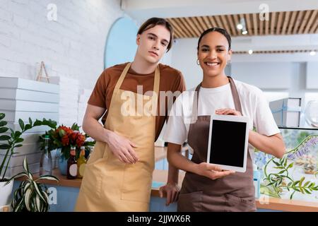 Un vendeur afro-américain positif montrant une tablette numérique près d'un collègue dans une boutique de produits sucrés Banque D'Images