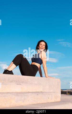 Corps complet de femme gaie dans des vêtements de sport assis sur un pas de béton pendant l'entraînement dans la rue le jour d'été ensoleillé dans la ville Banque D'Images