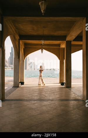 Femme marchant sous l'arche d'un bâtiment arabe traditionnel près de la mer tout en passant une journée d'été à Doha, au Qatar Banque D'Images