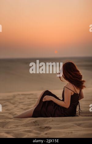 Vue latérale d'une femme anonyme aux longs cheveux rouges, assise sur du sable sec et admirant le ciel ensoleillé dans le désert près de Doha, Qatar Banque D'Images