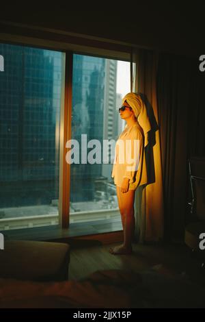Vue latérale de la jeune femme touristique barefooted en chemise blanche et lunettes de soleil avec une serviette sur la tête debout près de la fenêtre panoramique dans l'appartement moderne Banque D'Images