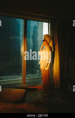 Vue latérale de la jeune femme touristique barefooted en chemise blanche et lunettes de soleil avec une serviette sur la tête debout près de la fenêtre panoramique dans l'appartement moderne Banque D'Images