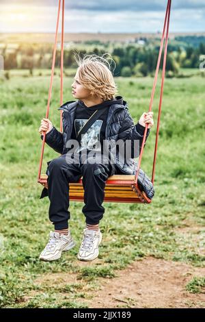 Un garçon d'école mignon avec de longs cheveux blonds manèges sur balançoire regardant de côté. Garçon aime la nature contre l'herbe verte et les arbres luxuriants lors d'une journée nuageux dans la campagne. N Banque D'Images