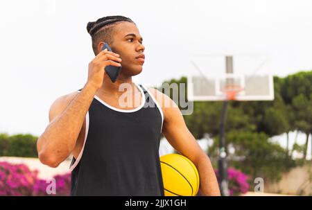 Jeune sportif hispanique avec ballon de basket-ball sur un appel téléphonique sur téléphone mobile tout en prenant une pause dans l'entraînement dans le parc regardant loin Banque D'Images