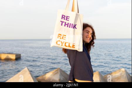 Jeune femme souriante avec le bras relevé tenant le sac tout en se tenant à la plage contre le ciel Banque D'Images
