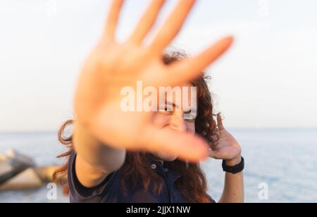 Portrait d'une jeune femme souriante montrant un geste d'arrêt tout en appréciant à la plage contre le ciel Banque D'Images
