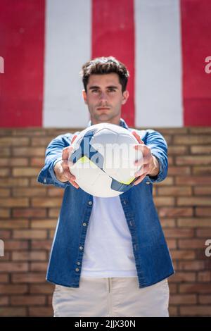 Portrait du jeune homme beau montrant le ballon de football en se tenant contre le mur en regardant l'appareil photo Banque D'Images