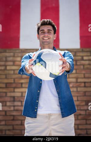 Portrait d'un beau jeune homme heureux montrant le ballon de football en se tenant contre le mur en regardant l'appareil photo Banque D'Images