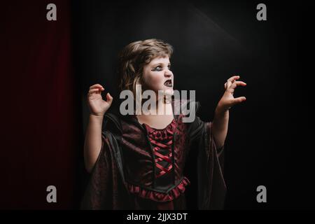 Petite fille créepy avec maquillage sombre dans le costume enchantress faisant peur visage et regardant loin sur fond noir pendant la fête d'Halloween Banque D'Images