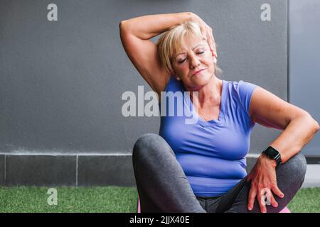 Posez la femme senior qui fait de l'exercice de cou assis avec les yeux fermés sur le tapis de yoga Banque D'Images