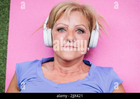 Vue de dessus gros plan d'une femme âgée qui écoute de la musique couchée sur un tapis de yoga regardant l'appareil photo Banque D'Images