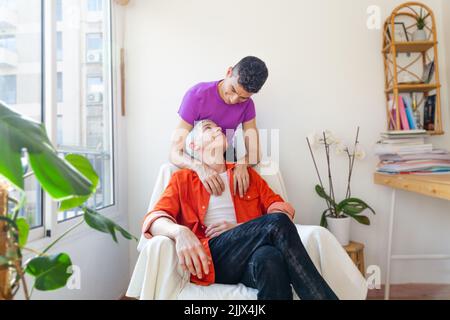 Portrait d'un couple gay souriant et aimant assis sur un fauteuil à la maison Banque D'Images