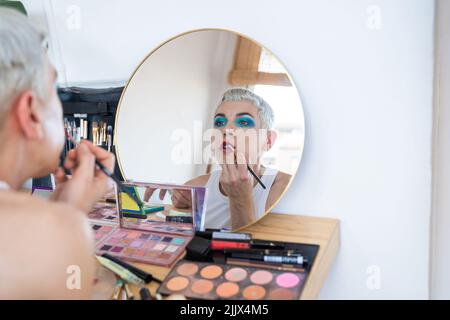 Jeunes transgenres regardant le miroir et appliquant le rouge à lèvres à la table de garniture Banque D'Images