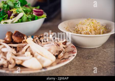 Assortiment de champignons crus dans une assiette placée sur la table près des pousses de haricots mung et de la salade fraîche dans la cuisine légère pendant le processus de cuisson Banque D'Images