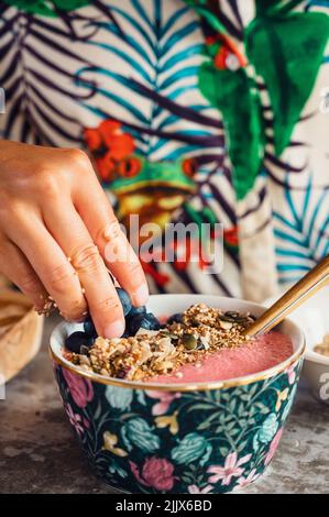 Récoltant des bleuets de mise anonymes dans un délicieux saladier à smoothie rose avec du granola pendant le petit déjeuner dans la cuisine à la maison Banque D'Images