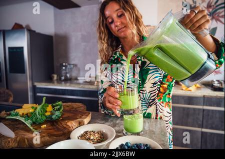Une femme attirante verse du smoothie vert sain dans un bol mélangeur dans un verre tout en se tenant près d'une table dans une cuisine légère à la maison Banque D'Images