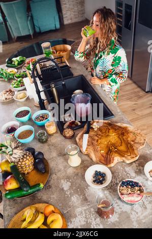 De la partie supérieure de la femme buvant un smoothie vert sain tout en se tenant à table avec des aliments et des repas variés dans la cuisine légère Banque D'Images