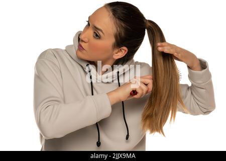 une jeune femme dans un survêtement gris brossant sa queue de poney sur un fond blanc dans le studio. Banque D'Images