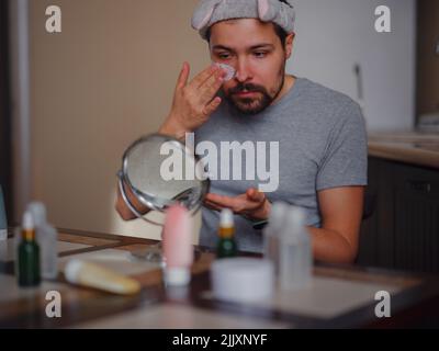 Homme de beauté concept. Caucasien gars vérifiant sa peau. Homme regardant dans le petit miroir tout en appliquant le masque de désintoxication de peau d'argile. Soin de la peau et soin masculin caucasien. Banque D'Images