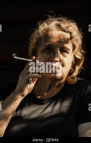 Portrait d'une femme âgée avec une cigarette en gros plan sur un fond naturel. Lumière du coucher du soleil sur le visage de l'aîné. Photo de haute qualité Banque D'Images