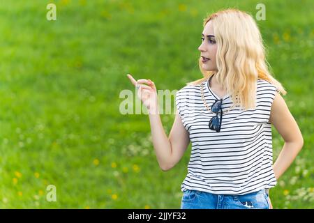 Blonde gitl caucasien pointant à côté et regardant sérieusement. Moyenne pousse vert herbe concept de publicité - espace de copie. Photo de haute qualité Banque D'Images