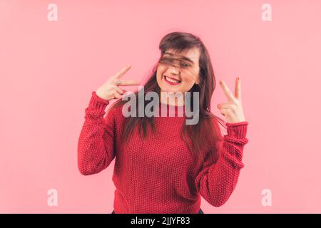 Femme brune caucasienne avec des cheveux coulant et un grand sourire portant un chandail rouge posant avec des signes de paix. Prise de vue en studio sur fond rose. Photo de haute qualité Banque D'Images