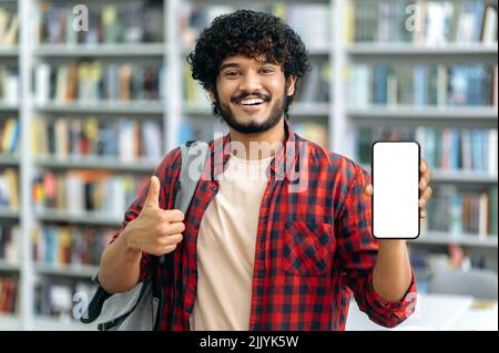 Gadgets, maquette, technologie sans fil. Un homme indien ou arabe positif, indépendant ou étudiant, montre un smartphone avec un écran maquette blanc et pointe le doigt dessus, regarde l'appareil photo, sourit avec joie Banque D'Images