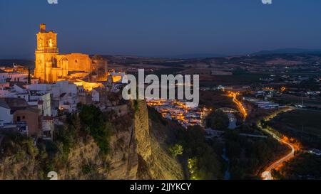 Iglesia de San Pedro à Arcos de la Frontera Espagne la nuit sur une falaise baignée de lumière jaune pendant l'heure bleue. Les lumières de la ville sont visibles ci-dessous. Banque D'Images