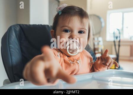 Portrait d'une jeune fille caucasienne assise sur une table à manger de bébé, regardant et pointant vers l'appareil photo. Alimentation saine, alimentation. Repas sain. Petit déjeuner sain Banque D'Images