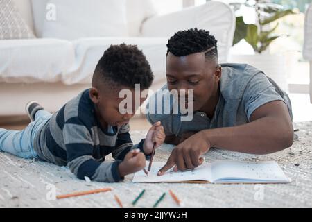 Un jeune père allongé sur le sol du salon et aidant son fils à dessiner. Banque D'Images