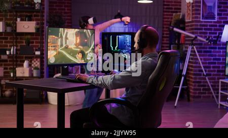Homme jouant en ligne multijoueur rpg d'action sur pc tandis que la fille de jeu combat dans le jeu de réalité virtuelle dans la salle de séjour. Le joueur diffuse le jeu de tir de première personne tandis que la femme utilise des lunettes vr. Banque D'Images