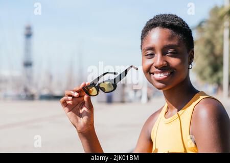 portrait d'une femme afro-américaine positive regardant l'appareil photo près du port. Elle porte un t-shirt jaune d'été et tient des lunettes de soleil jaunes Banque D'Images