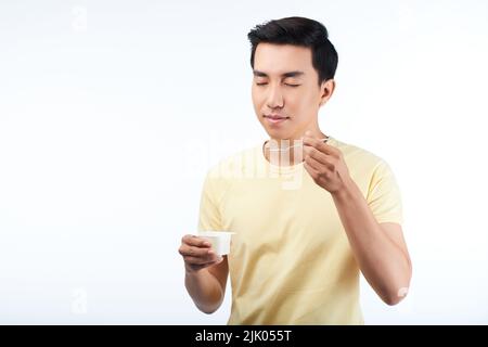 Portrait à la taille du beau asiatique avec les yeux fermés debout sur fond blanc et en appréciant le goût du yaourt Banque D'Images