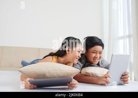 Frère et sœur gaies allongé sur un canapé et regardant des dessins animés sur une tablette Banque D'Images