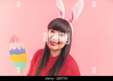 Charmante femme heureuse brune portant de mignons oreilles de lapin de Pâques et tenant un œuf de pâques en papier coloré sur un bâton, souriant à l'appareil photo et s'amusant. Photo de haute qualité Banque D'Images