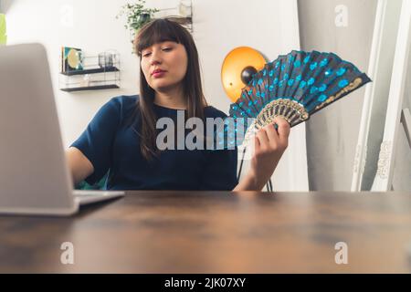 Une jeune femme freelance brune s'assoit à une table derrière son ordinateur portable à la maison et travaille à distance tout en utilisant un ventilateur à main en raison de la chaleur estivale. Photo de haute qualité Banque D'Images