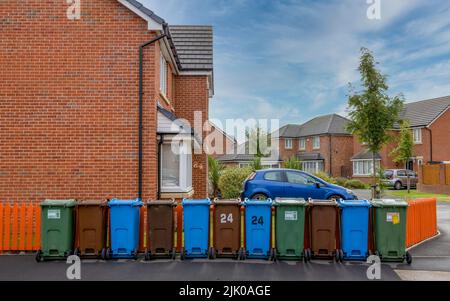 Une rangée de bennes à roulettes colorées pour le recyclage du plastique, du papier et des déchets de jardin, à côté d'une clôture orange et à l'extérieur d'une maison moderne. Banque D'Images