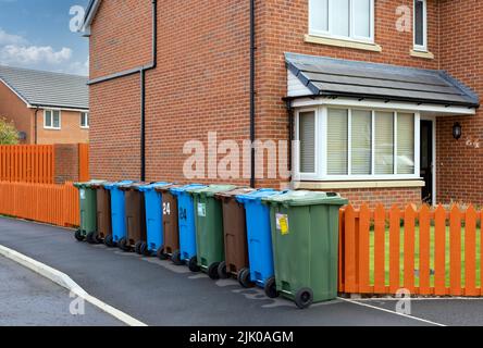 Une rangée de bennes à roulettes colorées pour le recyclage du plastique, du papier et des déchets de jardin, à côté d'une clôture orange et à l'extérieur d'une maison moderne. Banque D'Images