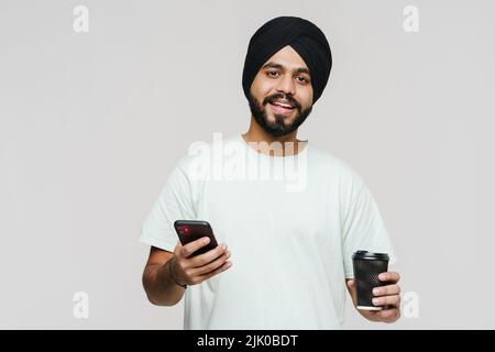 Homme sud-asiatique barbu utilisant un téléphone portable et buvant du café isolé sur un mur blanc Banque D'Images