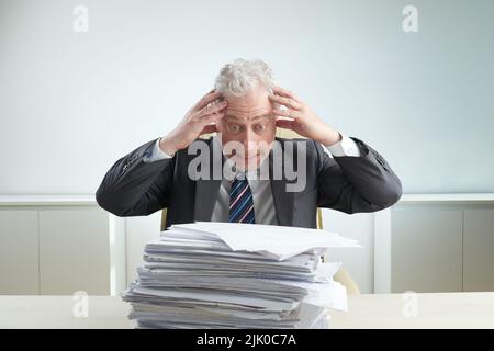 Homme d'affaires expérimenté stressé portant un costume tenant la tête et regardant une pile de documents avec colère tout en étant assis au bureau, portrait à la taille haute Banque D'Images