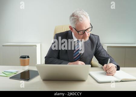 Homme d'affaires âgé concentré portant un costume classique et des lunettes de vue prenant les notes nécessaires tout en étant assis au bureau et en préparant pour les négociations importantes, portrait tourné Banque D'Images