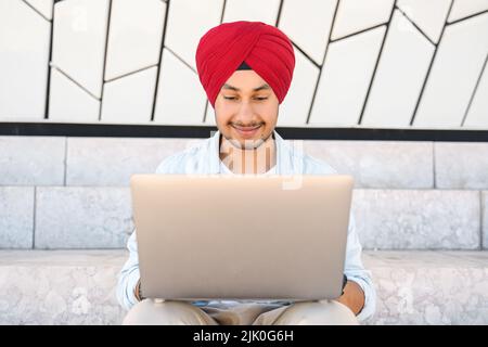 Homme indien gai portant un turban de tête de lit assis sur les marches à l'extérieur et utilisant un ordinateur portable. Inspiré heureux hindou homme entrepreneur dactylographiant sur le clavier et regarde l'écran avec plaisir sourire Banque D'Images
