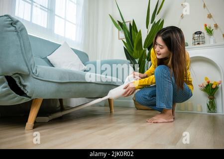 Femme de ménage nettoyage de la poussière sol sous canapé ou mobilier de canapé avec aspirateur, jeune femme asiatique heureuse avec accumulateur aspirateur à hom Banque D'Images