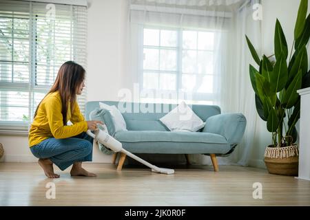 Femme de ménage nettoyage de la poussière sol sous canapé ou mobilier de canapé avec aspirateur, jeune femme asiatique heureuse avec accumulateur aspirateur à hom Banque D'Images