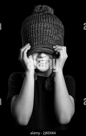 Une photo verticale en niveaux de gris d'un petit garçon heureux couvrant ses yeux avec un chapeau d'hiver doux et souriant Banque D'Images