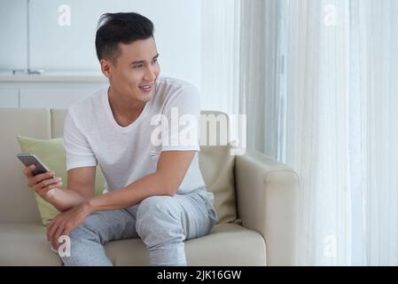 Portrait d'un jeune homme souriant avec un smartphone assis sur son canapé Banque D'Images