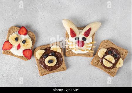 Quatre toasts doux pour les enfants en forme de poussin, ours, lapin et singe, avec fraises, banane, fromage à la crème, chocolat et flocons de noix de coco sur b gris Banque D'Images