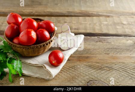 Tomates mûres rouges dans un bol en osier avec une branche de persil et d'ail sur fond de bois. Le concept de la cuisine végétarienne. Orientation horizontale. Banque D'Images